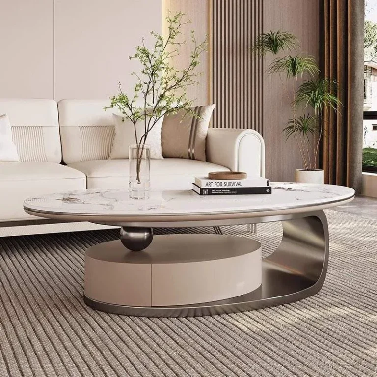 Oval ivory sintered-stone table on a curved grey metal base with a taupe drum and a ball pivot, in front of a cream sofa