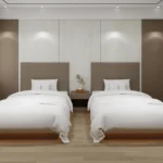 Two beds side by side with tall taupe padded headboards, a small bedside table between and wood-tone wall columns