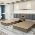 Two taupe platform bed bases side by side with gray padded headboards and a small nightstand between
