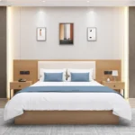 Made-up bed with a cream padded headboard, blue cushions and runner, and two one-drawer nightstands with phone and lamp