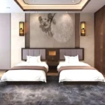 Twin beds with gray-mauve padded headboards, a shared nightstand with a lamp, wall sconces and a leaf-pattern wall mural