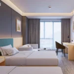 Bed with a teal-blue padded headboard on a light-wood wall panel, a gray sofa with a round table and a desk with chair