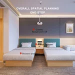 Twin beds with teal-blue headboards, a shared nightstand with a lamp and a gray sofa at the right