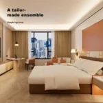 Wide room view with the bed, a sofa with orange cushions at the window, a round table with chair and the orange wall panel