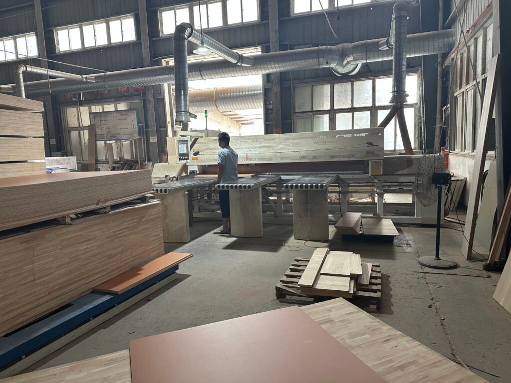 Operator at a woodworking machine beside stacked furniture panels in the workshop.