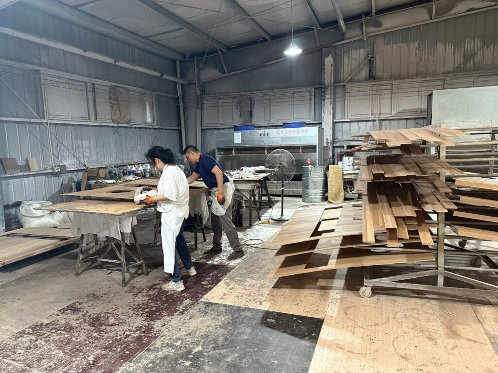 Workers preparing wooden panels beside racks of furniture components.