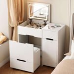 White foldaway vanity with the lid up and a lit LED mirror, a side cabinet and a pull-out seat, beside a bed