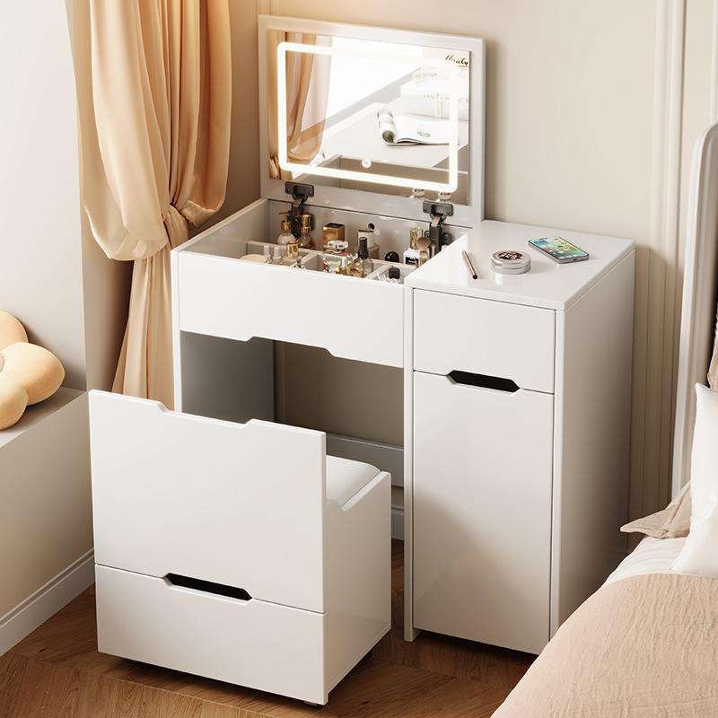 White foldaway vanity with the lid up and a lit LED mirror, a side cabinet and a pull-out seat, beside a bed