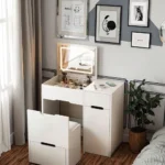 White foldaway vanity with a flip-up LED mirror and the seat pulled out, in front of framed pictures and a plant