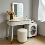 White vanity desk with a lit mirror on tapered legs, a side cabinet with an oval window and a round fluted stool