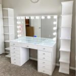 White vanity with a bulb-lit mirror, a glass-covered desktop, two drawer banks and two shelf towers, on a carpet
