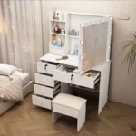 White vanity with drawers pulled open and a bulb-framed mirror swung out, beside a padded stool
