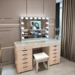 Beige vanity with a lit bulb mirror, a glass-covered top, drawer banks and a stool with a mirrored frame