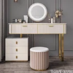 Cream-white fluted vanity with a round LED mirror, a three-drawer cabinet and a pink stool, against a gray paneled wall
