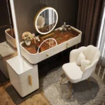Cream vanity with an amber glass top, a gold-framed round LED mirror, a side cabinet and a scalloped chair, beside curtains