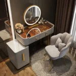 Gray vanity with an amber glass top, a gold-framed round LED mirror and a gray scalloped chair